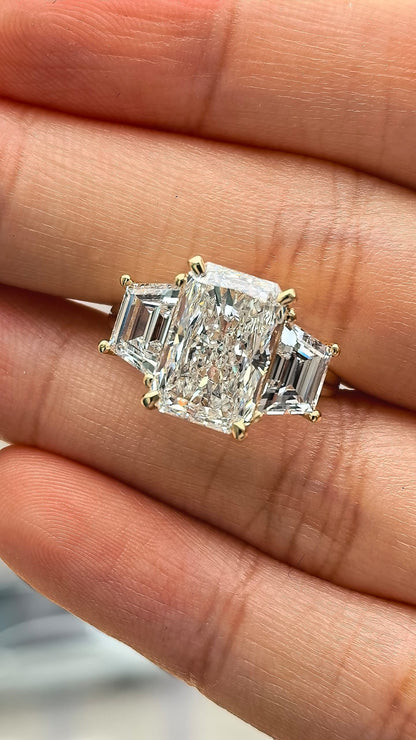 A close-up of a diamond ring held on a person's fingers. The ring features a large rectangular emerald-cut center stone with two smaller baguette-cut side stones, set in a yellow gold band.