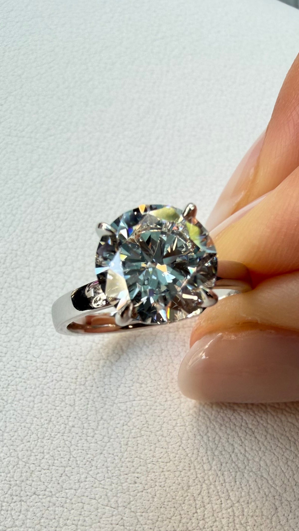 A silver solitaire ring with a large round brilliant-cut gemstone held between fingers against a textured off-white background.