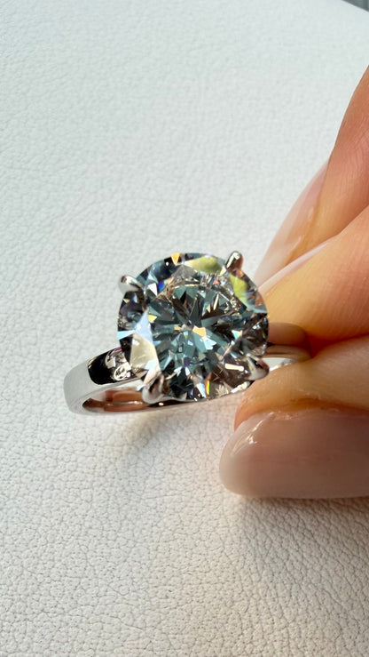 A silver solitaire ring with a large round brilliant-cut gemstone held between fingers against a textured off-white background.