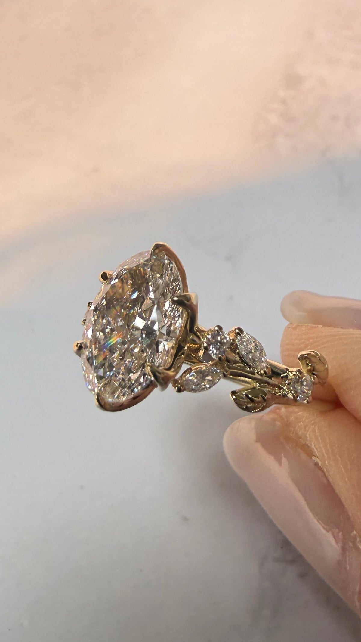 A close-up of a hand holding a gold ring with a large round clear gemstone and smaller accent stones on a light marble-like surface.