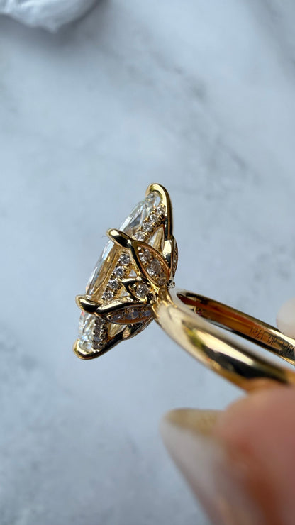 A close-up of a yellow gold ring with a marquise-cut diamond center stone and smaller diamonds along the shoulders, held by a hand against a light marble background.