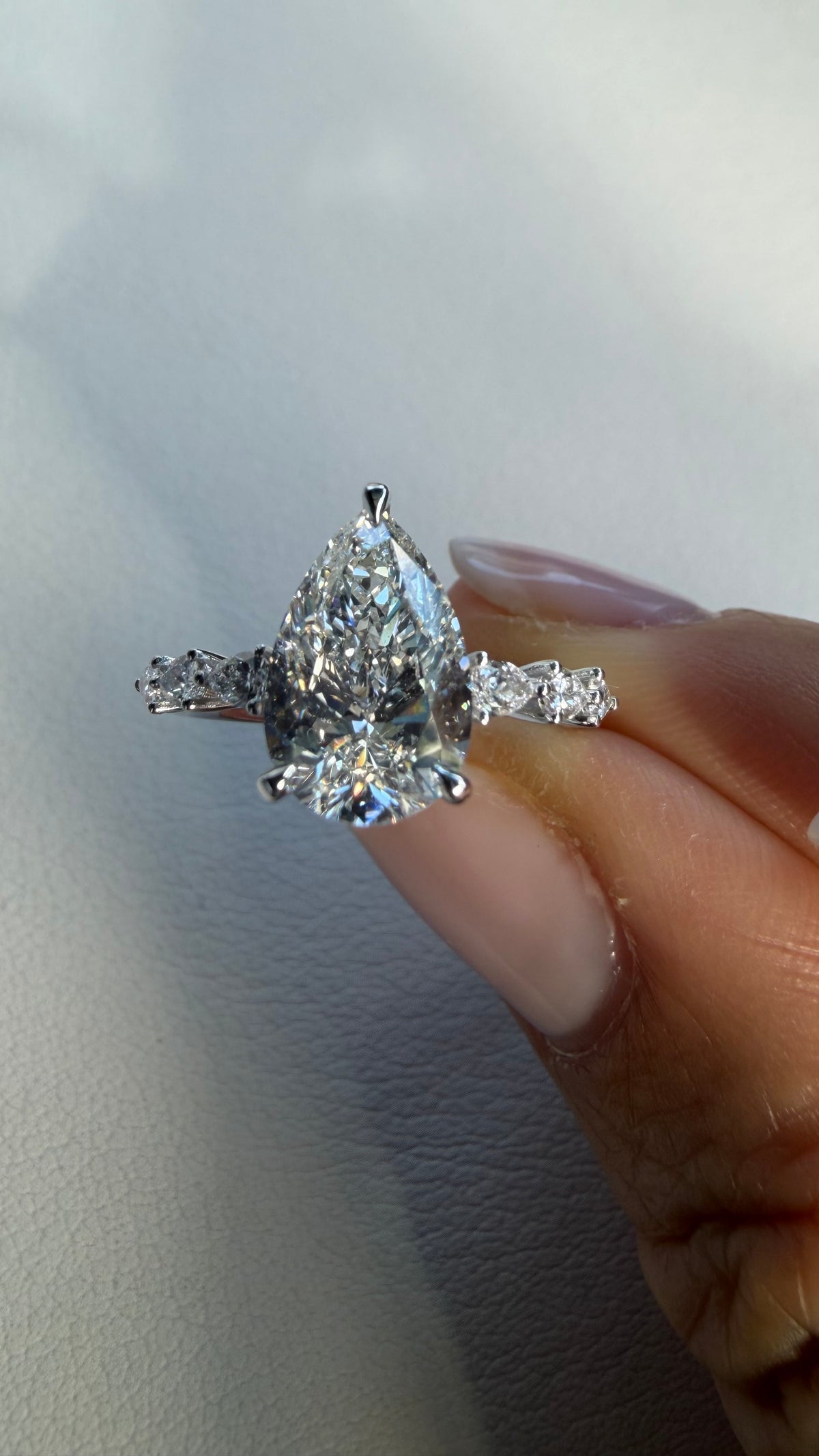 A pear-cut diamond engagement ring with diamond accents on the band, held between fingers against a light gray background.