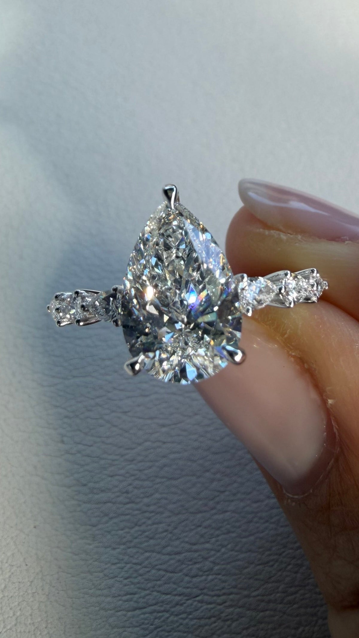 A pear-cut diamond ring with baguette side stones on a silver-toned band, held between fingers against a light gray textured background.